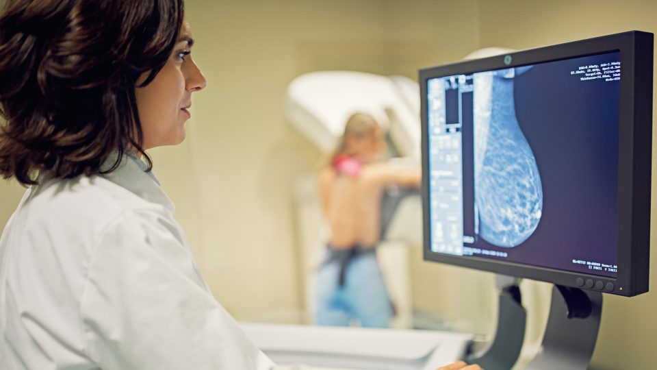 Doctor Examines Real-Time Breast Imaging Result on Computer Screen, While Patient Is Imaged in Background