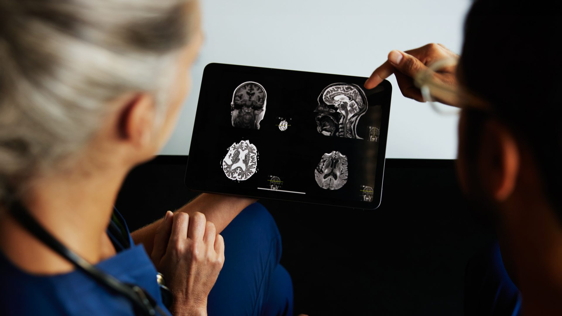 One healthcare provider holding a tablet with brain scan images while another healthcare provider points to one of the images